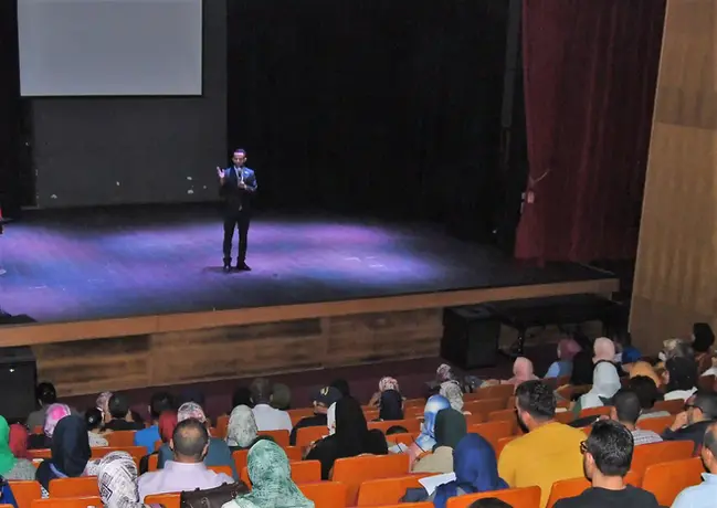 رجل يقف على المسرح يتحدث إلى جمهور جالس، إضاءة مسرح، عرض تقديمي