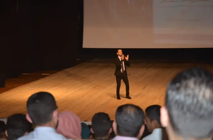 رجل يتحدث على المسرح أمام الجمهور، مع عرض تقديمي في الخلفية. مؤسسة صُناع القرار