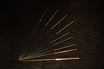A photograph of nine silver acupuncture needles, four longer than the other five. They're arranged into a quarter-circle with their tips touching and handles evenly spaced against a dark brown wood grain background.