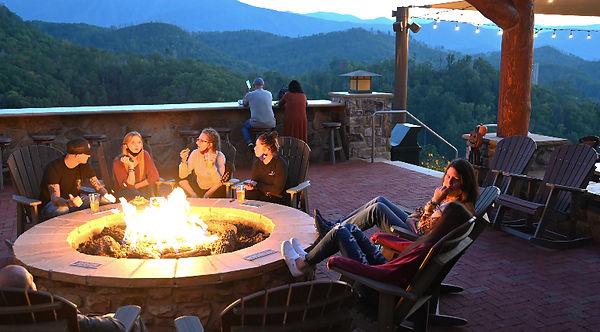People gather around a large fire table at Anakeesta Attraction in Gatlinburg Tennessee, with the Smoky Mountains behind them in the evening
