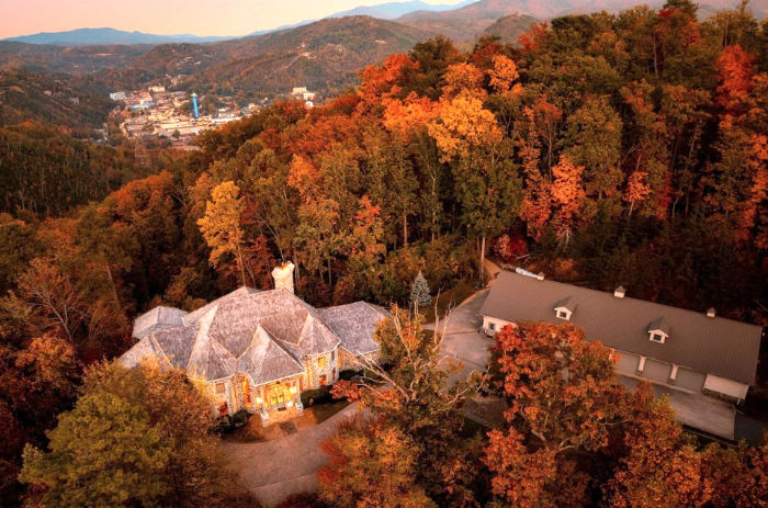Aerial view of a house and long building in autumn forest, with orange-red trees, mountains, and the distant town of Gatlinburg Tennessee below.