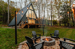 A Frame Cabin in front of a forested mountain, with a flat grassy area that has a stone firepit wih a warm fire burning, surrounded by chairs.