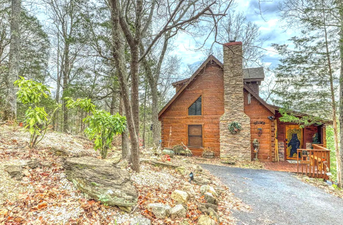 Rustic log cabin with stone chimney on a gravel drive in a wooded forest, surrounded by bare trees and spring greenery.