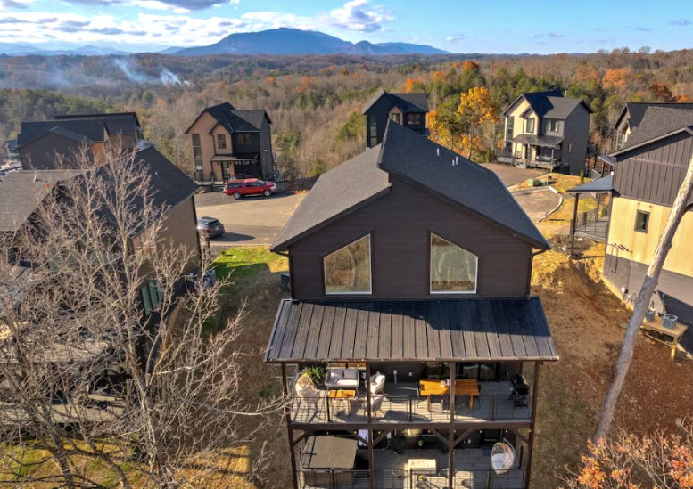 Cluster of modern cabins with dark roofs in autumn hills, a red SUV on a drive, and distant blue mountains under a clear sky.