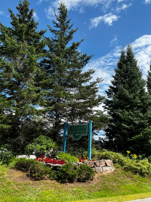 Sign surrounded by flowers and tall pine trees in background, reads "Aiglon Tremblant" and is on road to Aiglon Tremblant condos