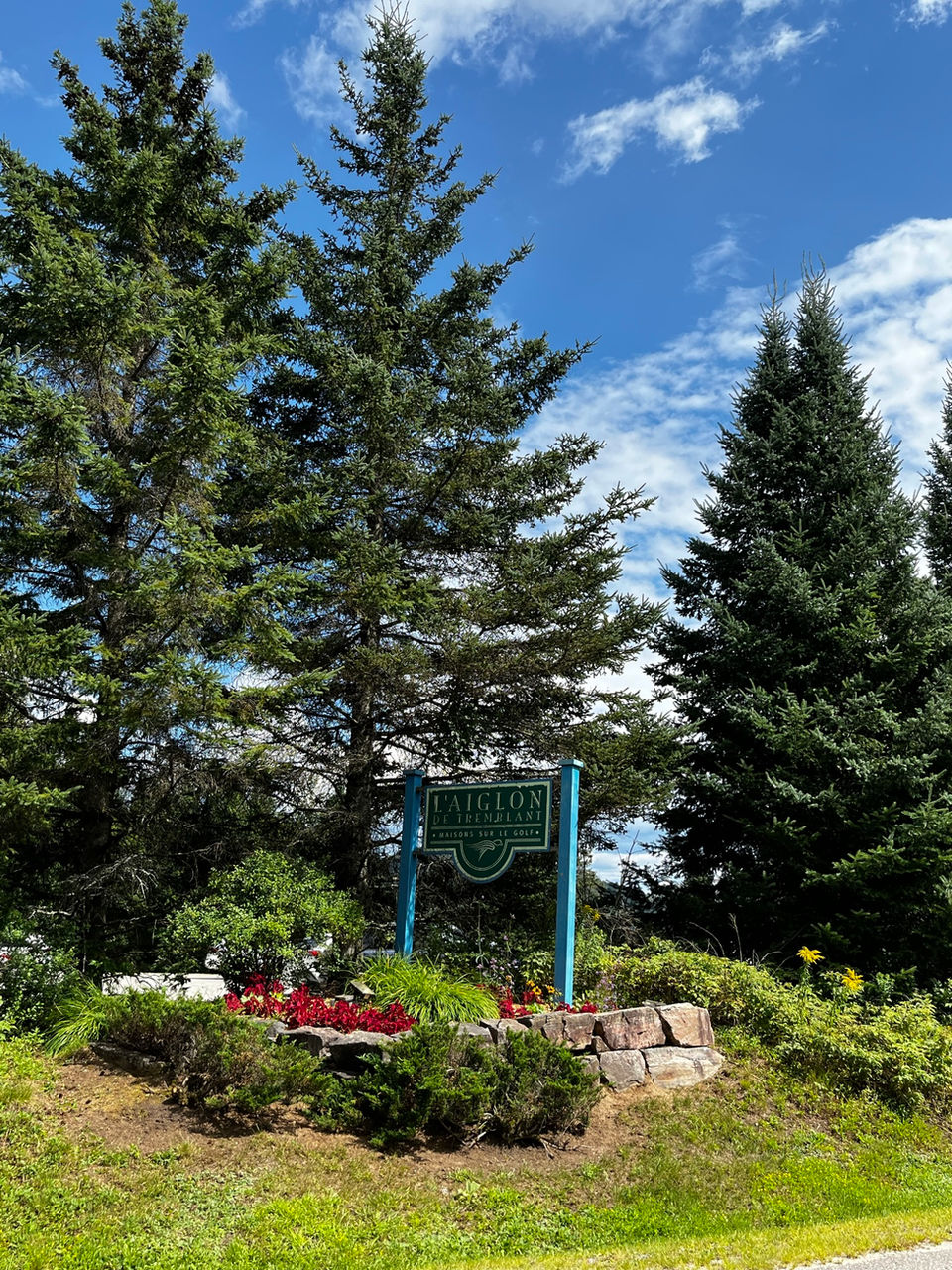 Sign surrounded by flowers and tall pine trees in background, reads "Aiglon Tremblant" and is on road to Aiglon Tremblant condos