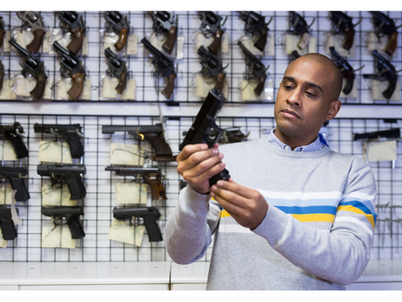 A person examines a handgun in a gun shop. Background displays multiple firearms. The person wears a grey sweater with colorful stripes.