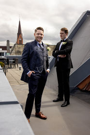 To menn i bryllupsantrekk på takterrasse; en smiler.