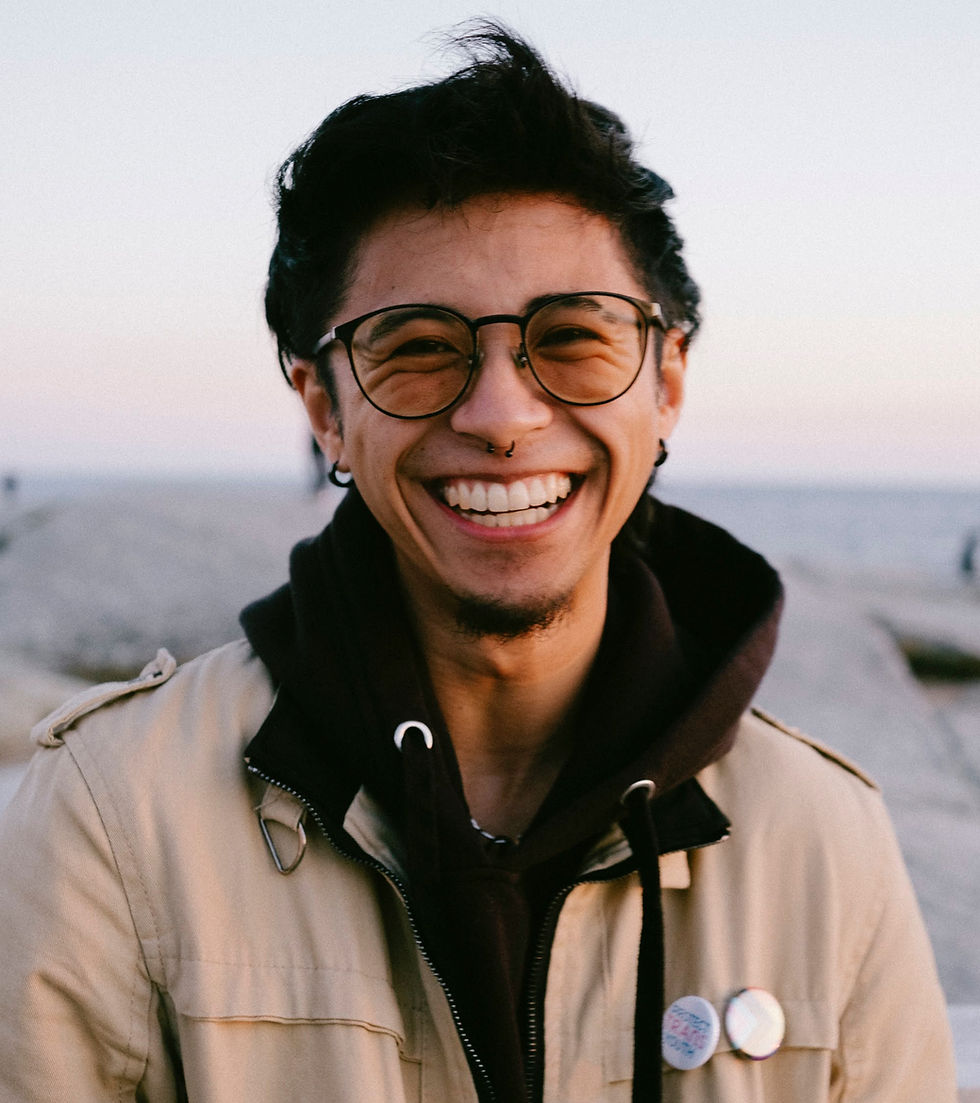 Cal smiling, standing in front of the shore. They’re wearing orange-tinted glasses, a black hoodie, and a beige jacket with white buttons on the chest pocket.