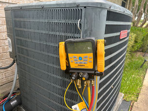 Fieldpiece gauge connected to a Goodman outdoor AC condenser beside a brick wall, with colored hoses and lawn in back.