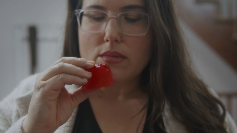 The mother holds a red ice cube with the fingertips of her right hand. She peers through her clear-rimmed glasses at the ice cube.