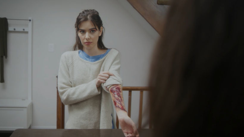 An over mom's shoulder view of Sandra standing on the opposite side of the kitchen island. She has her left sleeve pulled up to exposed several gashes covering her inner forearm, of varying age. She looks at her mother with concern for her reaction.