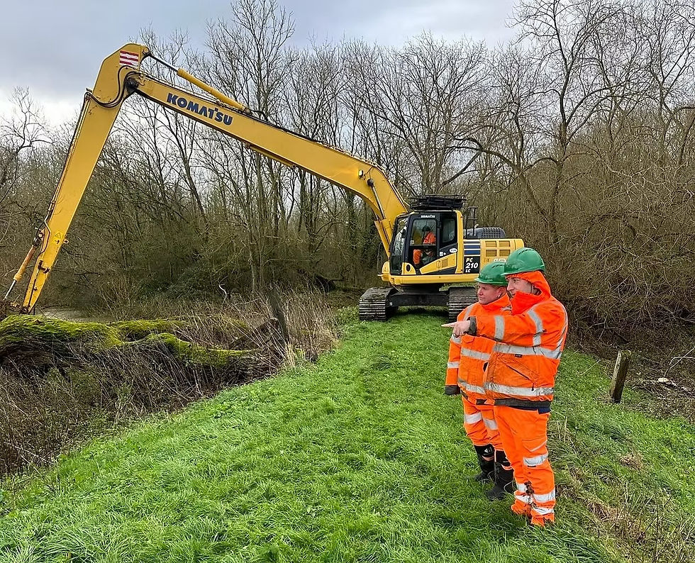 Two workers in orange safety gear point beside a Komatsu excavator on a grassy ditch under leafless trees.
