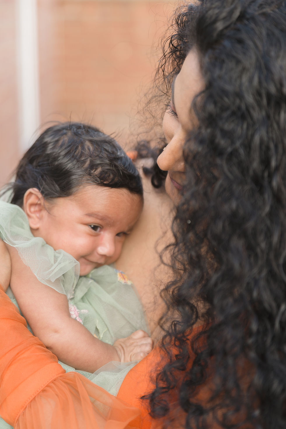 Mother cuddles sleeping baby in pale green dress, close-up against a soft peach background.
