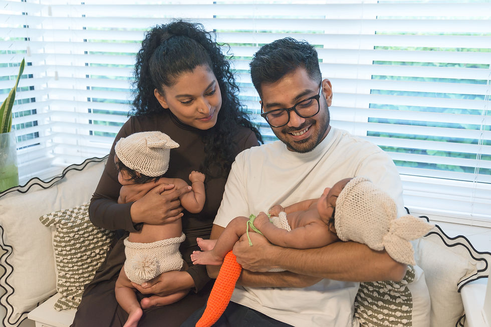 Parents smile while holding two sleeping babies in knit hats on a couch beside bright window blinds.
