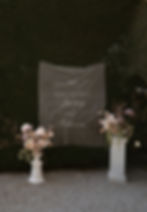 Large embroidered wedding day sign displayed between tall floral plinths in a garden setting.