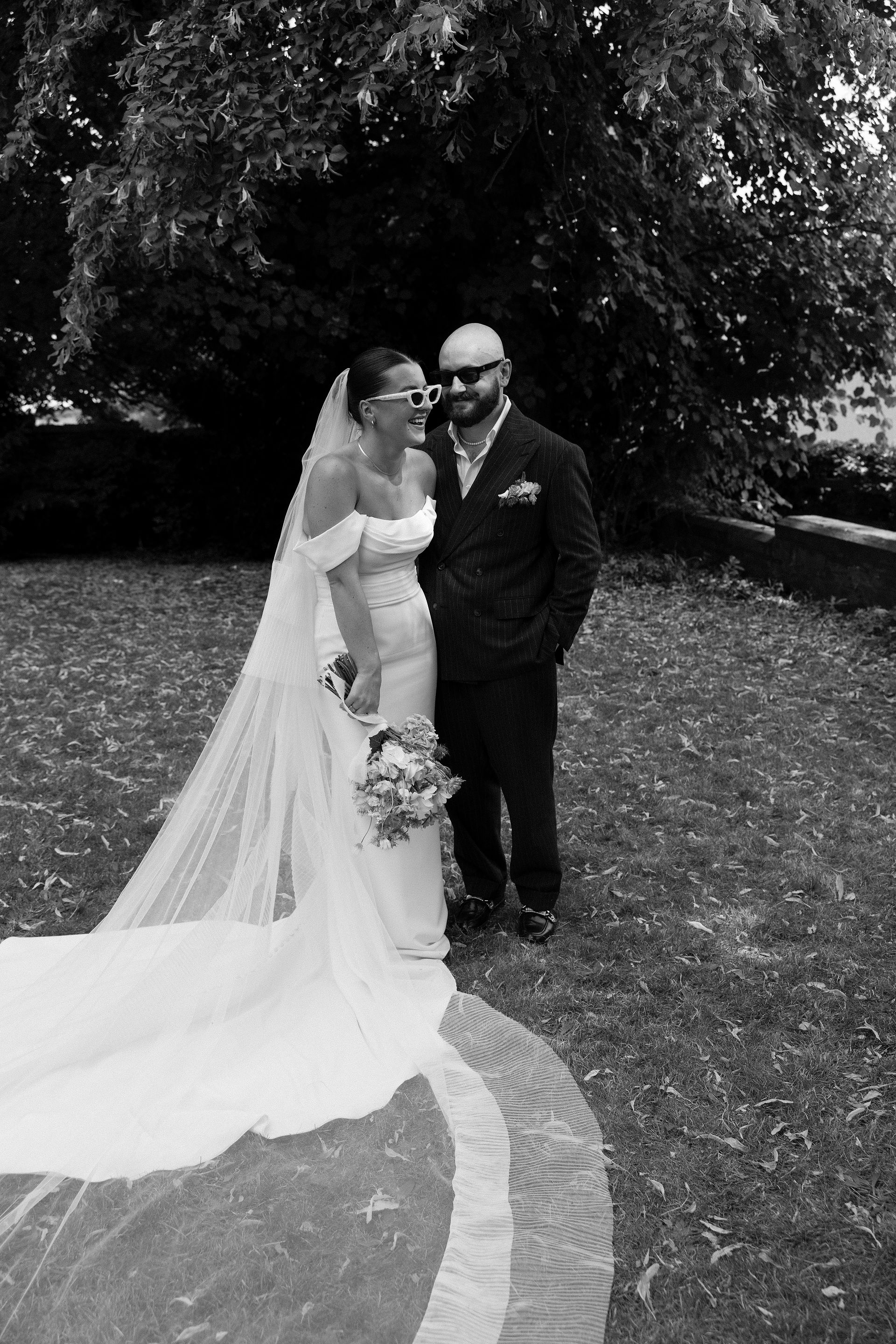 A bride in an off-the-shoulder gown wears a flowing pleated edge veil, standing beside her groom in a garden setting.