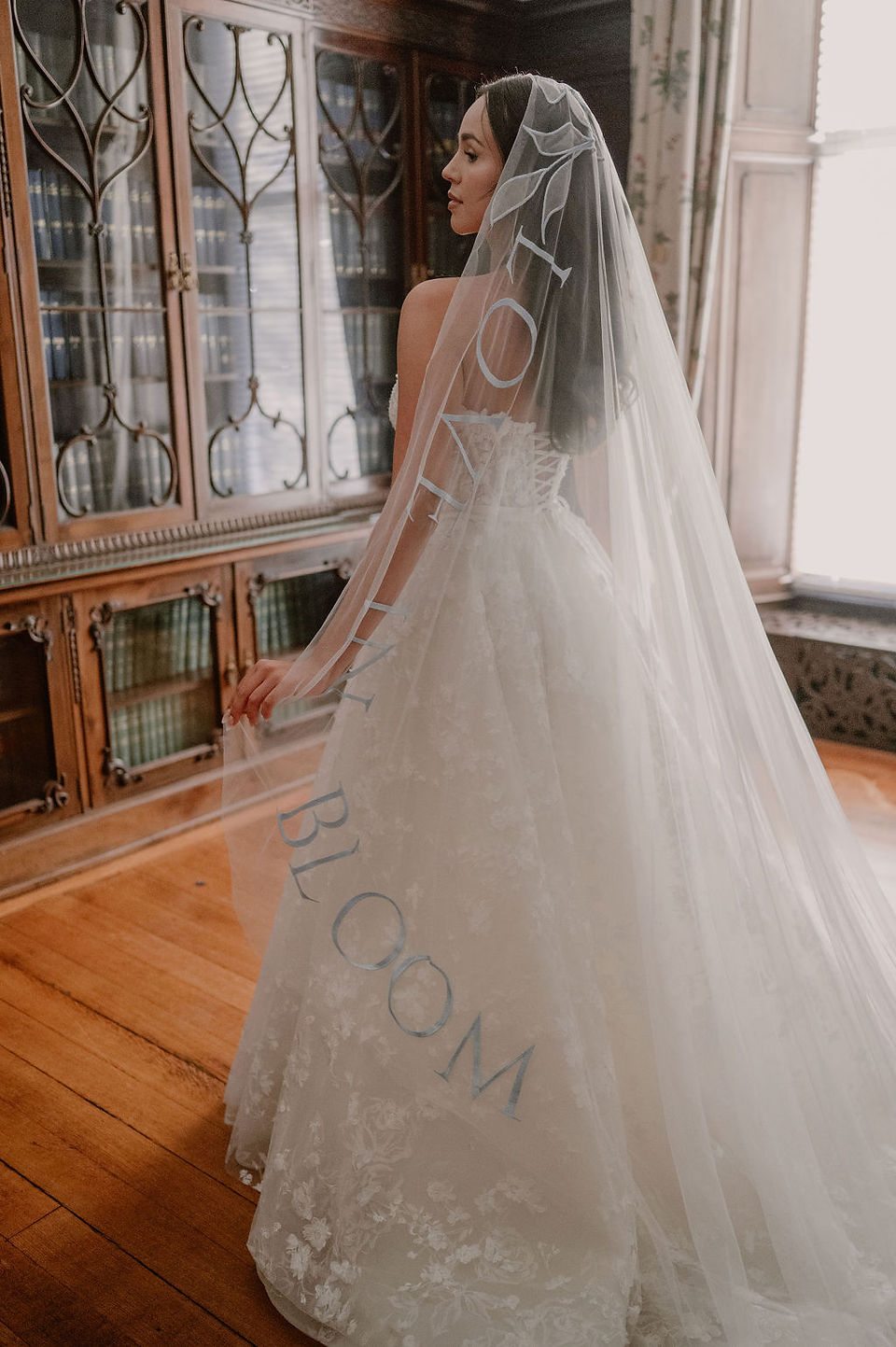 Back view of a bride wearing a coloured wedding veil with bespoke dusky blue script embroidery reading “Let Love Bloom.”