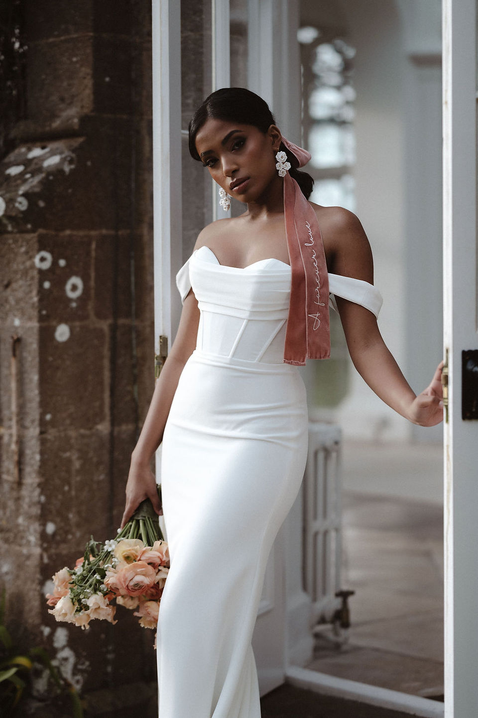A bride in an off-the-shoulder white gown wears The Limited Edition Velvet Ribbon in peach, embroidered with ivory script, tied in her hair.