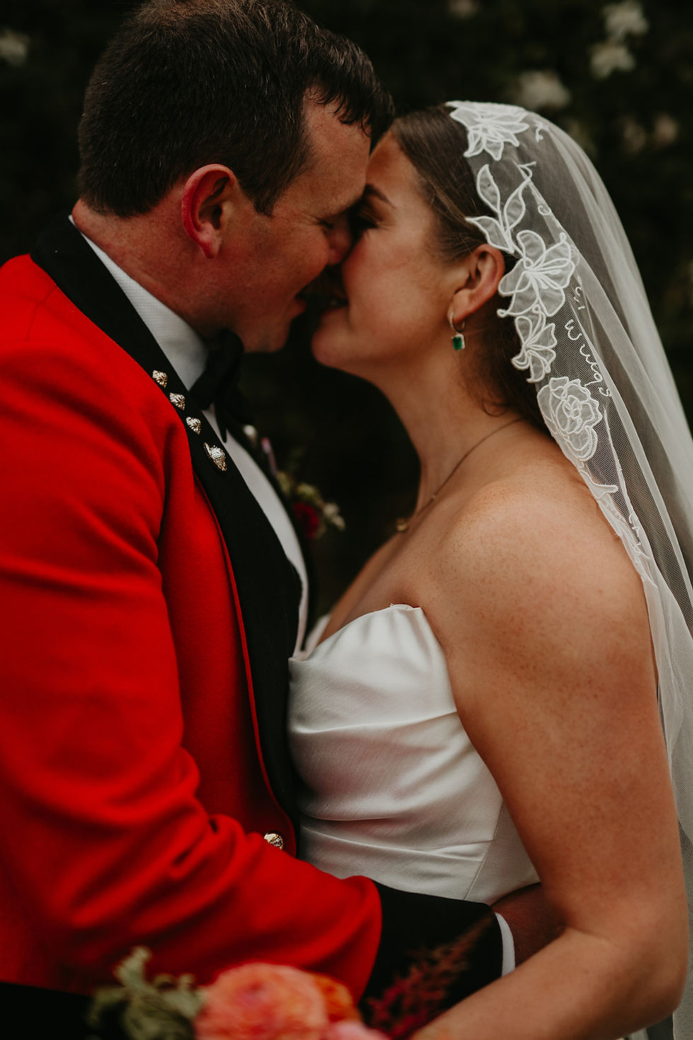 Military groom in red jacket kissing bride wearing floral embroidered mantilla veil at naval wedding.