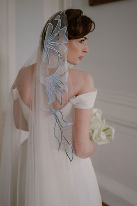 Back view of a bride holding white calla lilies, showcasing the bold blue floral embroidery on her personalised mantilla wedding veil.
