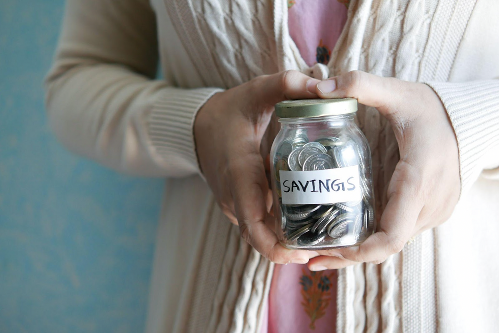 An elderly woman holds a jar that says “savings” with coins inside.