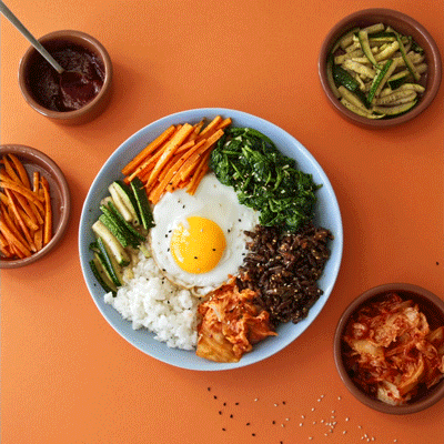 The gif shows a bowl of Korean bibimbap with rice, carrots, cucumbers, kimchi, spinach, egg and crickets with gochujang sauce. On the side in small dishes, there are carrots, cucumbers, kimchi and sauce. A hand sprinkles bibimbap with spring onion. Edible insects.