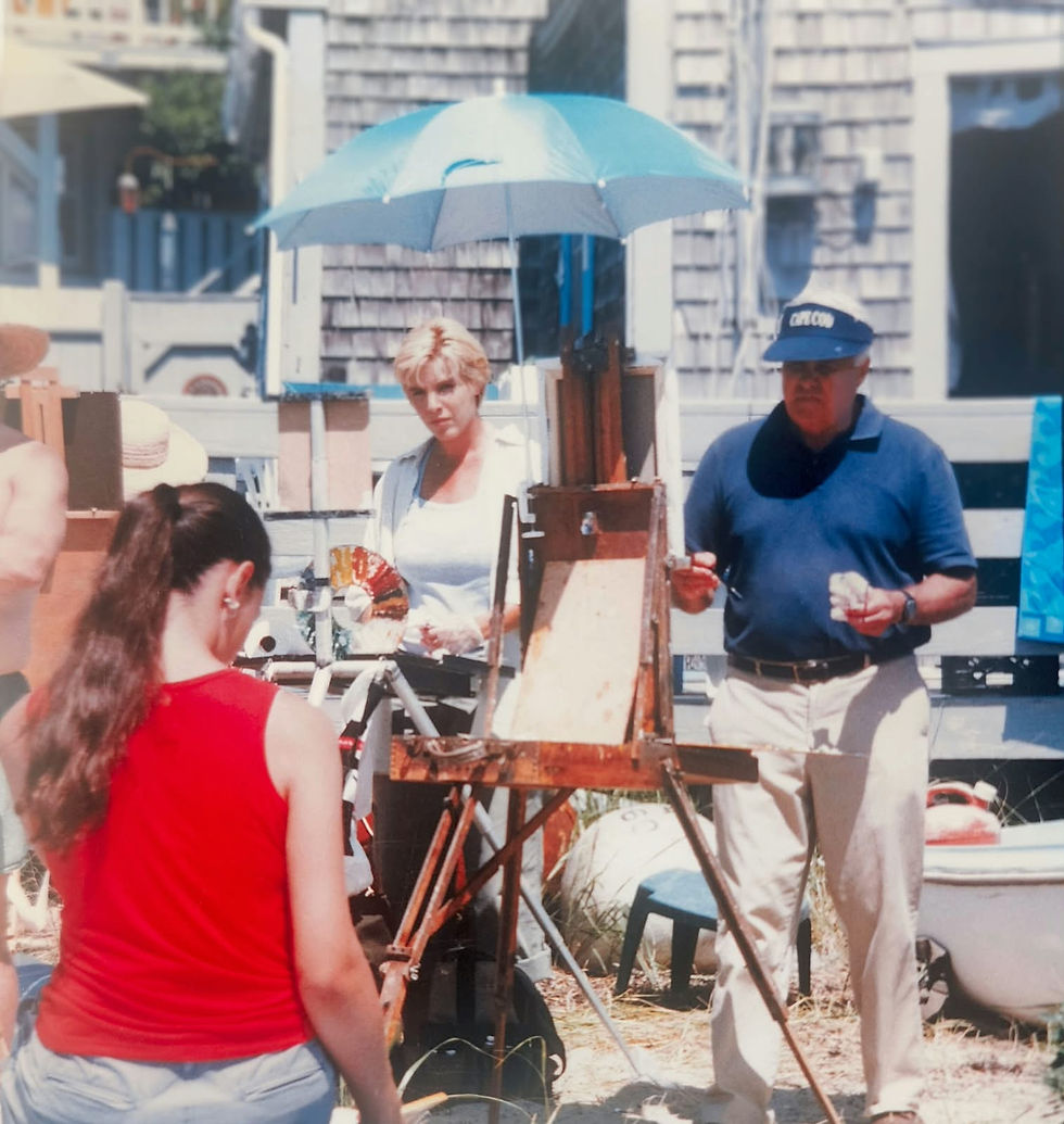 Carole Gray-Weihman and Cedric Egeli painting on the beach in Provincetownl, with a blue umbrella overhead and houses behind; sunny, focused on painting Sarah, the model dressed in red.