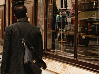 A person in a dark coat and messenger bag walks past a shop with cane displays. Reflections seen in windows. Sign reads "James Smith & Sons."