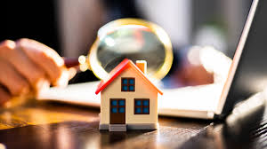 A toy house on a desk with a magnifying glass hovering above, next to a laptop. Hand in background. Warm lighting, focused study atmosphere.