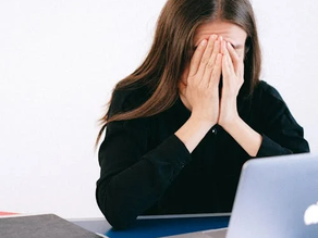 A person in a black shirt covers their face with hands at a desk with a laptop, papers, and glasses. The mood appears stressed in a bright room.