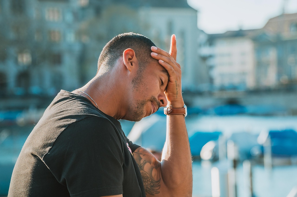 Man outdoors by the water, eyes closed and hand on forehead, looking stressed against a blurred city waterfront. illustrating hyperarousal