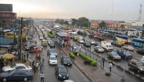 Busy city street in Surulere with cars and buses in traffic. Pedestrians walk on sidewalks. Overcast sky. Buildings and greenery in the background.