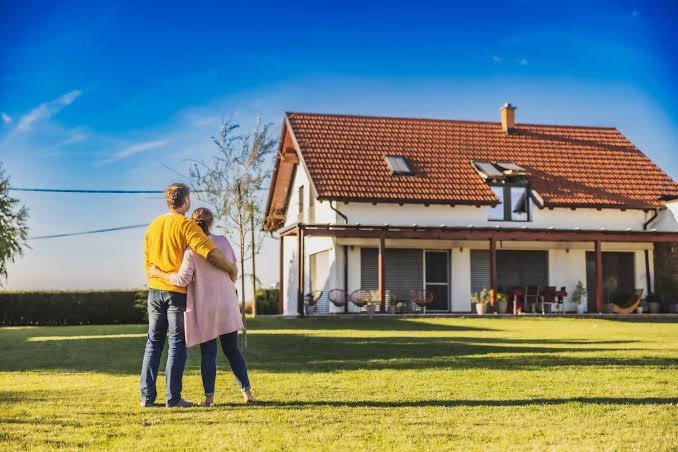 Couple embraces in a sunny lawn facing a red-roofed house with a porch under a clear blue sky.