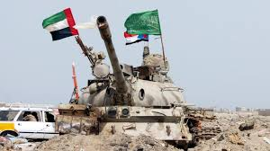 A tank with flags on its turret stands amidst rubble. UAE, Saudi, and other flags are visible. A car is in the background under a clear sky. Illustrating the Yemen war.