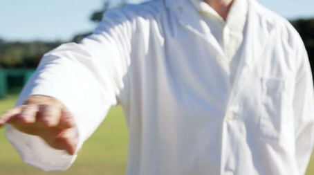 Person in a white lab coat extends an arm forward. Outdoor setting with blurred greenery. Bright, sunny day.