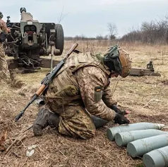 Soldiers in camouflage prepare artillery shells in a grassy field. One soldier kneels, focused on loading. The sky is overcast. Ukraine war