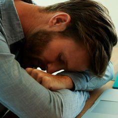 Man in blue shirt resting his head on folded arms at a desk with a laptop and turquoise folder, conveying exhaustion and burnout.
