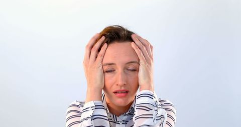 Woman in a striped shirt holding her head with both hands, eyes closed, appears stressed or exhausted, against a plain background.
