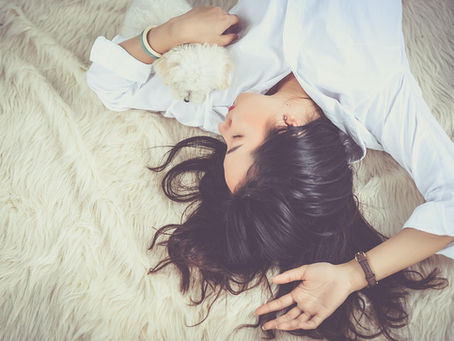 A woman in a white shirt lies on a fluffy cream carpet, peacefully cuddling a small white dog. Illustrating how sleep affects your mental health