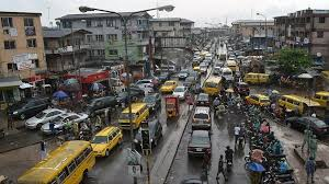 Busy urban street in Yaba with yellow buses, motorcycles, and pedestrians under cloudy skies. Buildings line the road, creating a lively, chaotic scene.