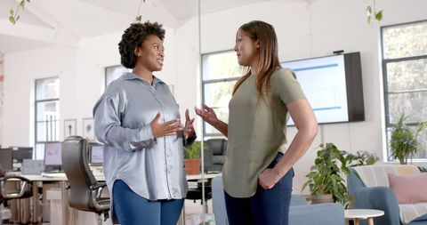 Two women chatting in a bright modern office with desks, plants, and a wall screen, appearing engaged and friendly.
