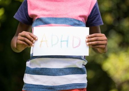 Child holds a sign with "ADHD" in multicolored letters. Wears a striped shirt. Background is a blurred green outdoor setting.