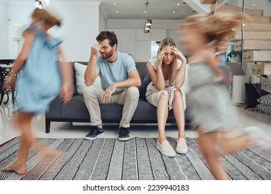 An exhausted mother and father sit on a couch looking worn out while their two young children energetically run around the living room.