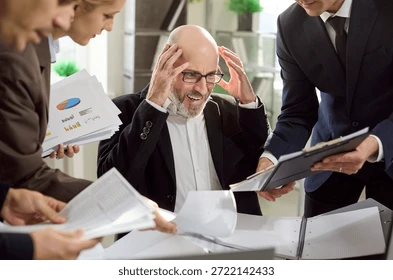 Business meeting with four people. An older man looks frustrated, holding his head. Papers and charts are scattered on the table.
