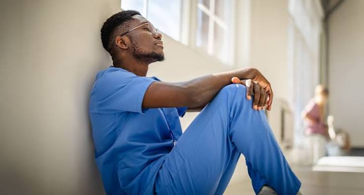 Man in blue scrubs sits against a wall in a bright hallway, resting with knees up and a thoughtful, tired expression.