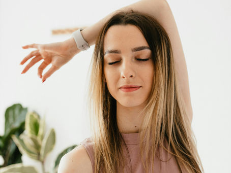 Woman in a pink top stands with eyes closed, arm raised, in a bright room with a plant. Peaceful expression, illustrating recovery