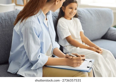 Woman with clipboard talks to smiling girl on a gray sofa in a bright living room; Shutterstock.com watermark below.