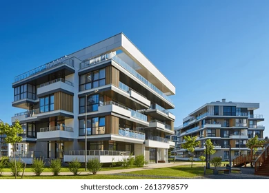 Modern apartment buildings with balconies, set against a clear blue sky. Greenery surrounds the structures, evoking a serene atmosphere.