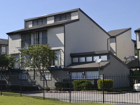 Modern townhouse with gray siding, trees in front, black fence, and satellite dishes. Clear blue sky in the background. Quiet, sunny setting.